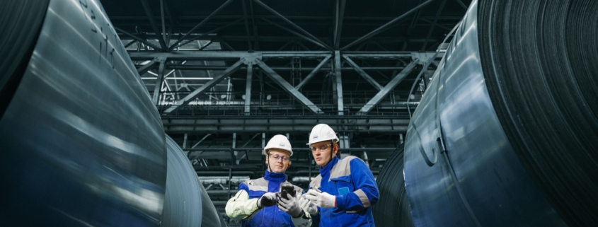 Two manufacturing workers inspecting steel coils at an industrial facility, illustrating in-house production and make-or-buy decisions